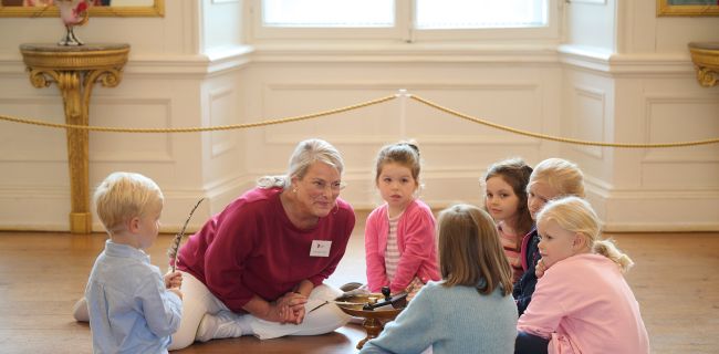 Eine Gruppe Kinder sitzt mit einer Museumsführerin auf dem Fußboden im Schloss