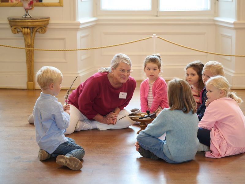 Eine Gruppe Kinder sitzt mit einer Museumsführerin auf dem Fußboden im Schloss